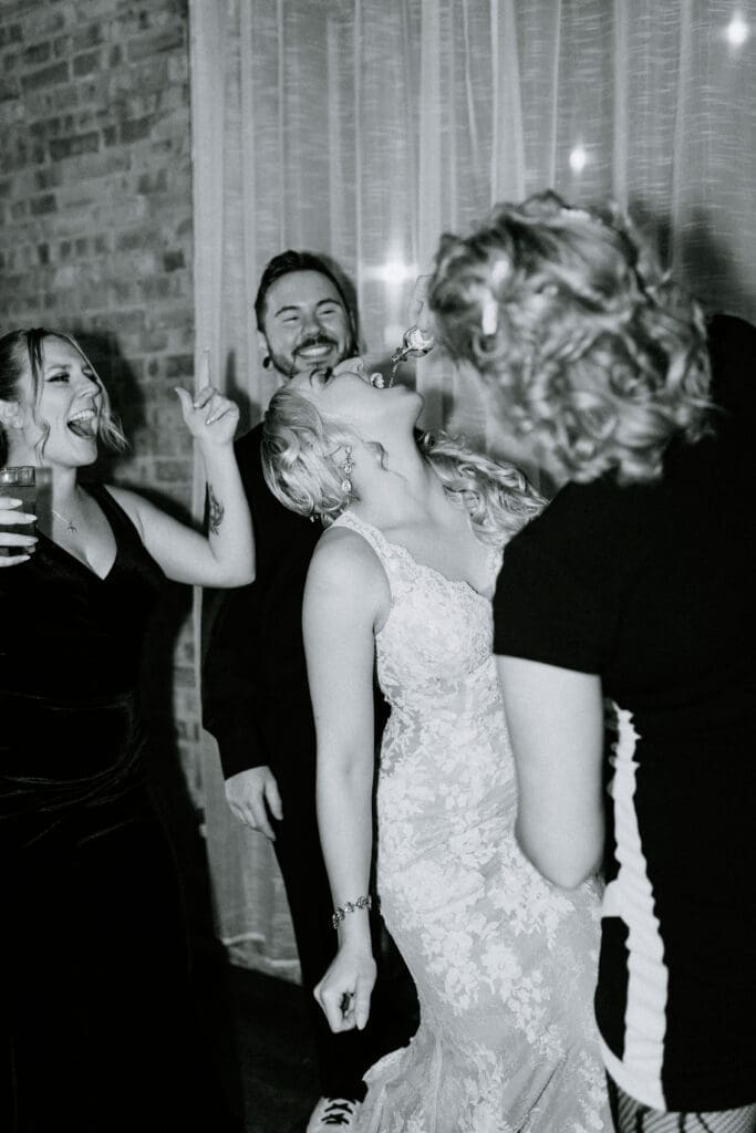 Bride takes a celebratory shot from her maid of honor as the dance floor opens while her groom and bridesmaid cheer and laugh beside her during the wedding reception.